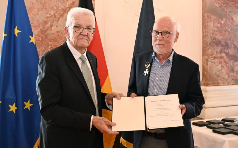 Zwei &auml;ltere M&auml;nner mit wei&szlig;em Haar mit einem Urkunden-Dokument. Ein Mann tr&auml;gt eine Medaille am Revers. Beide blicken in die Kamera. Im Hintergrund sind die Europa-, Deutschland- und Baden-W&uuml;rttemberg-Fahne zu sehen.