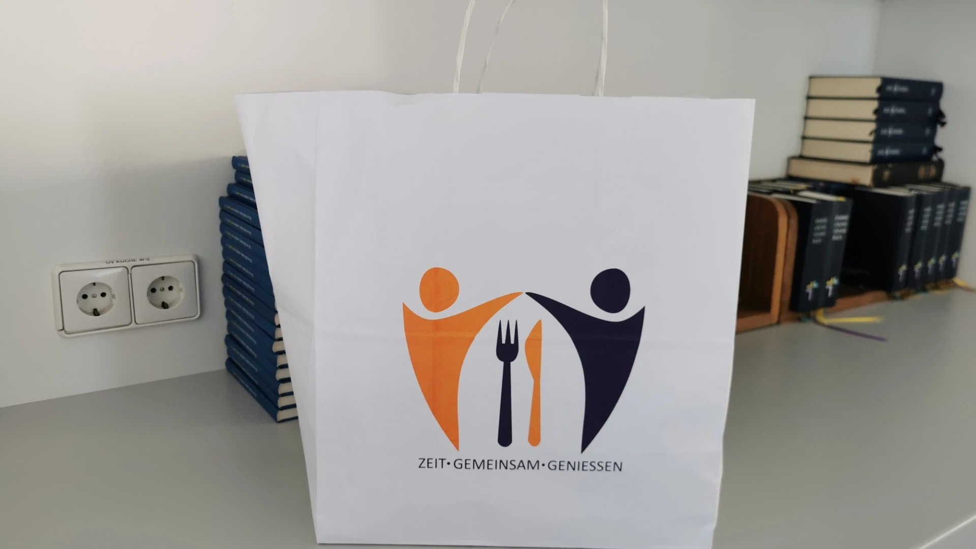 Wei&szlig;e Papiert&uuml;te mit orangefarbenem und dunkelblauem Logo, das zwei stilisierte Figuren und ein Besteck symbolisiert, mit dem Slogan "Zeit-gemeinsam-Geniessen" in einem Regal stehend vor aufgestapelten B&uuml;chern und Steckdosen.