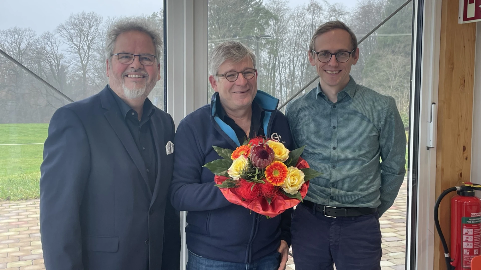 Drei Personen stehen in einem Raum vor einem gro&szlig;en Glasfenster mit Blick ins Gr&uuml;ne und l&auml;cheln in die Kamera. Der Mann in der Mitte h&auml;lt einen Blumenstrau&szlig; in der Hand. Rechts ist ein Feuerl&ouml;scher zu sehen.