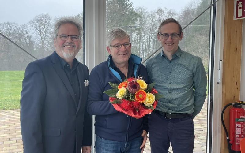 Drei Personen stehen in einem Raum vor einem gro&szlig;en Glasfenster mit Blick ins Gr&uuml;ne und l&auml;cheln in die Kamera. Der Mann in der Mitte h&auml;lt einen Blumenstrau&szlig; in der Hand. Rechts ist ein Feuerl&ouml;scher zu sehen.