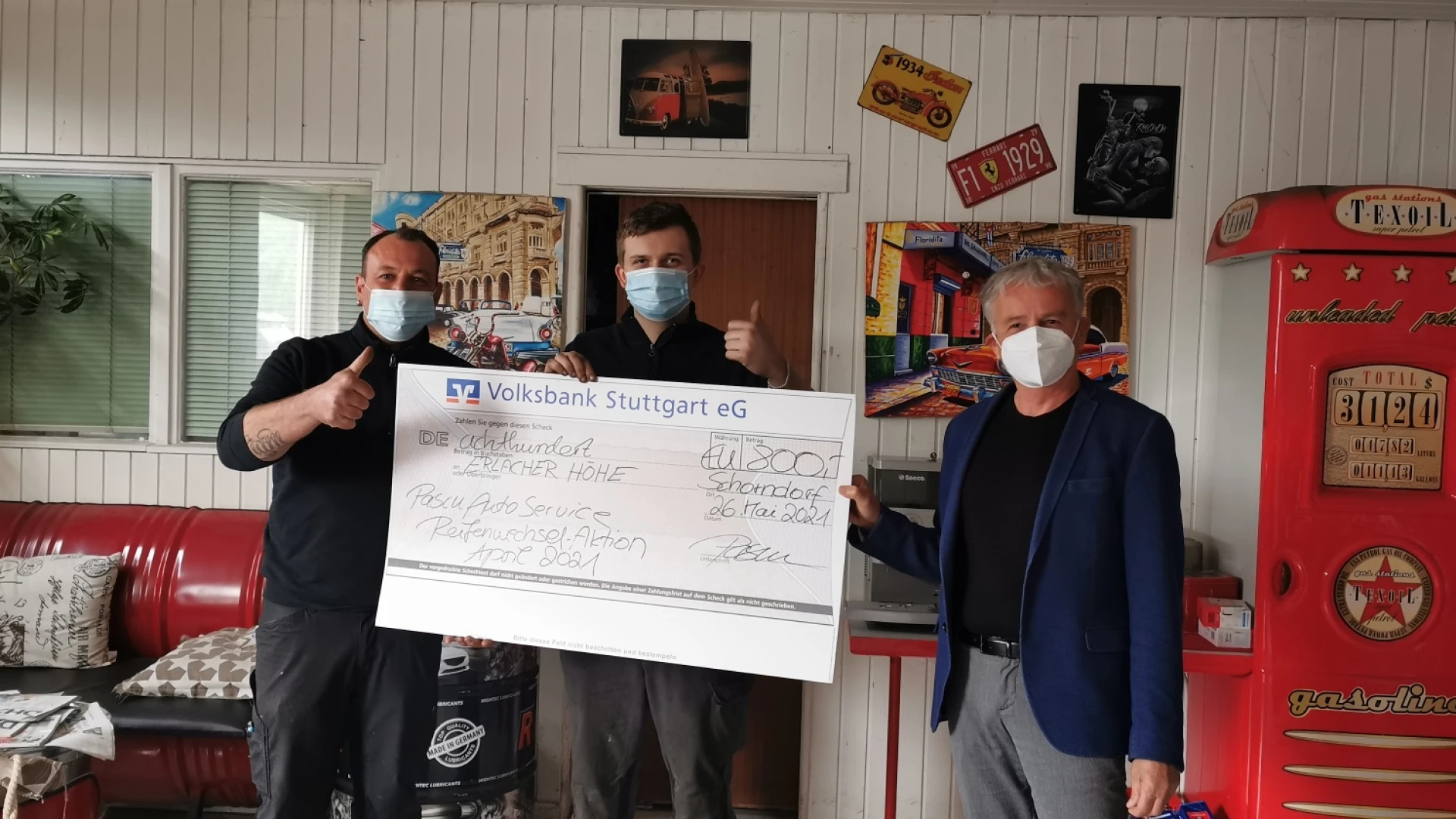 Drei M&auml;nner mit Mund-Nasenschutz stehen in einem Raum und halten gemeinsam einen Scheck in der Hand. Zwei der M&auml;nner zeigen mit dem Damen nach oben. Im Hintergrund ist eine rote Couch, Wandbilder, Fenster und ein wei&szlig;es Holzpaneel zu sehen.