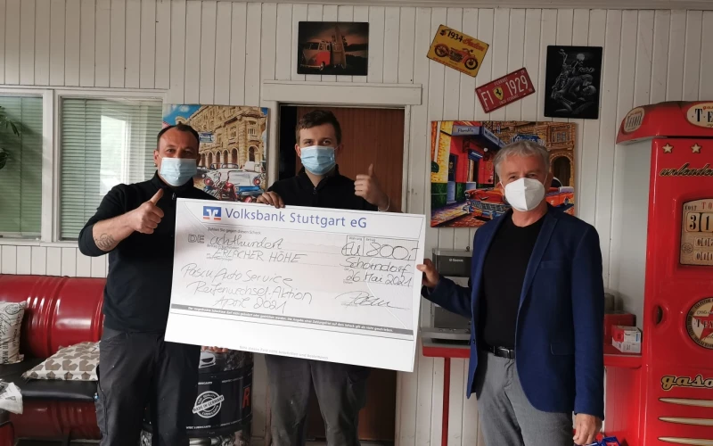 Drei M&auml;nner mit Mund-Nasenschutz stehen in einem Raum und halten gemeinsam einen Scheck in der Hand. Zwei der M&auml;nner zeigen mit dem Damen nach oben. Im Hintergrund ist eine rote Couch, Wandbilder, Fenster und ein wei&szlig;es Holzpaneel zu sehen.