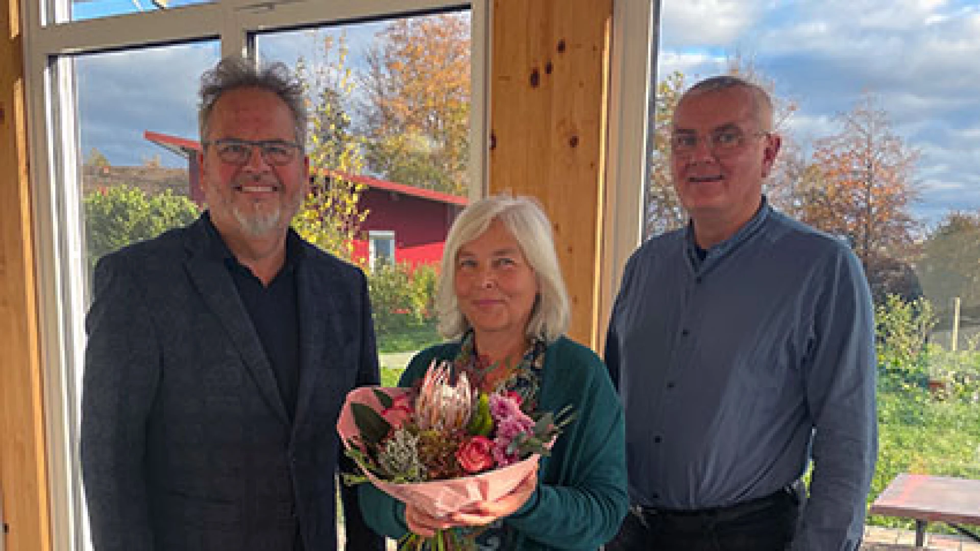Drei Personen stehen in einem Raum vor einer gro&szlig;en Fensterfront, die den Blick nach drau&szlig;en freigibt. Die Frau in der Mitte h&auml;lt einen Blumenstrau&szlig; in den H&auml;nden.