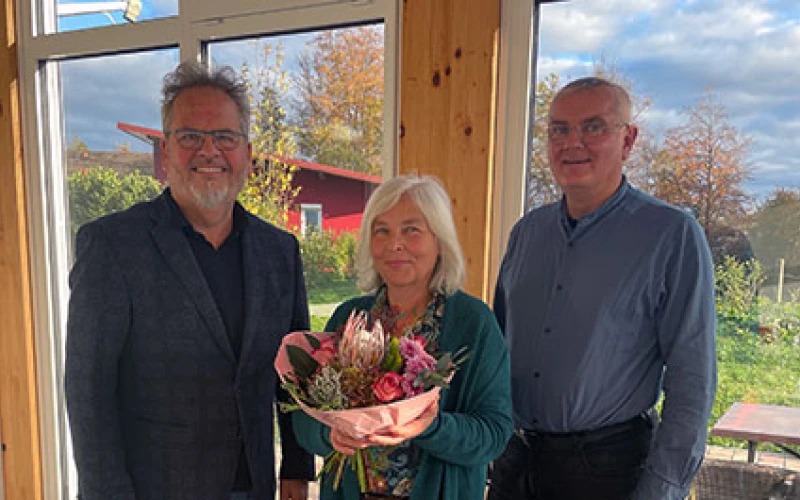 Drei Personen stehen in einem Raum vor einer gro&szlig;en Fensterfront, die den Blick nach drau&szlig;en freigibt. Die Frau in der Mitte h&auml;lt einen Blumenstrau&szlig; in den H&auml;nden.