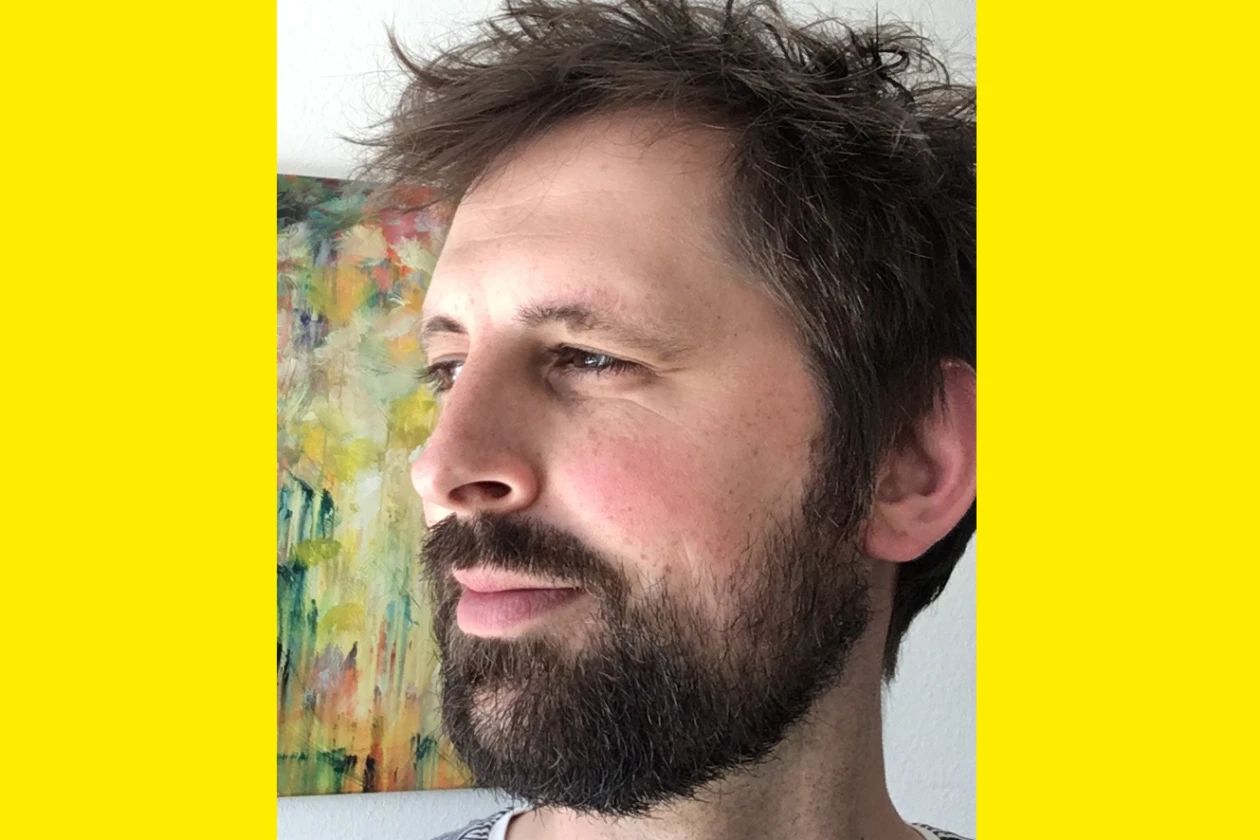 Der Kopf eines j&uuml;ngeren Mannes mit dunklem Bart und zerzausten Haaren ist seitlich in der Kamera zu sehen. Im Hintergrund ein Gem&auml;lde.