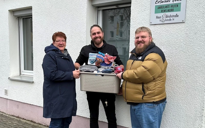 Eine Frau und zwei M&auml;nner stehen vor einem Geb&auml;ude mit einem Schild "Erlacher H&ouml;he" und l&auml;cheln in die Kamera. Ein Mann h&auml;lt einen Korb mit verschiedenen Utensilien in der Hand. Es scheint sich um eine Sachspende zu handeln.