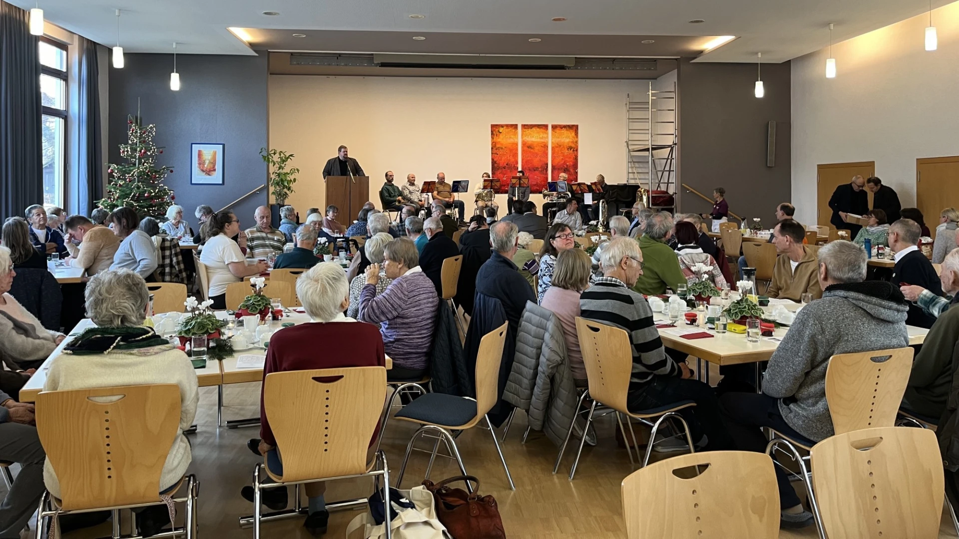 Gro&szlig;er Veranstaltungsraum mit mehreren eingedeckten Tischen, an denen Menschen sitzen. An der Wand h&auml;ngen rote Kunstwerke, und Deckenleuchten sorgen f&uuml;r eine helle Atmosph&auml;re. Eine Person steht vorne am Podium und scheint zu sprechen.