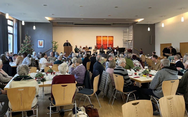 Gro&szlig;er Veranstaltungsraum mit mehreren eingedeckten Tischen, an denen Menschen sitzen. An der Wand h&auml;ngen rote Kunstwerke, und Deckenleuchten sorgen f&uuml;r eine helle Atmosph&auml;re. Eine Person steht vorne am Podium und scheint zu sprechen.