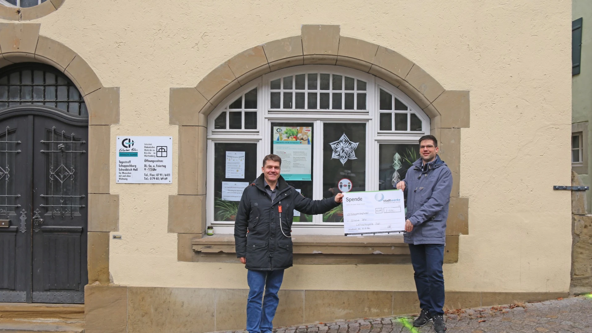 Zwei M&auml;nner stehen vor einem Geb&auml;ude mit einem gro&szlig;en bogenf&ouml;rmigen Fenster, blicken in die Kamera und halten gemeinsam einen Spendenscheck in der Hand.