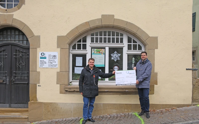 Zwei M&auml;nner stehen vor einem Geb&auml;ude mit einem gro&szlig;en bogenf&ouml;rmigen Fenster, blicken in die Kamera und halten gemeinsam einen Spendenscheck in der Hand.