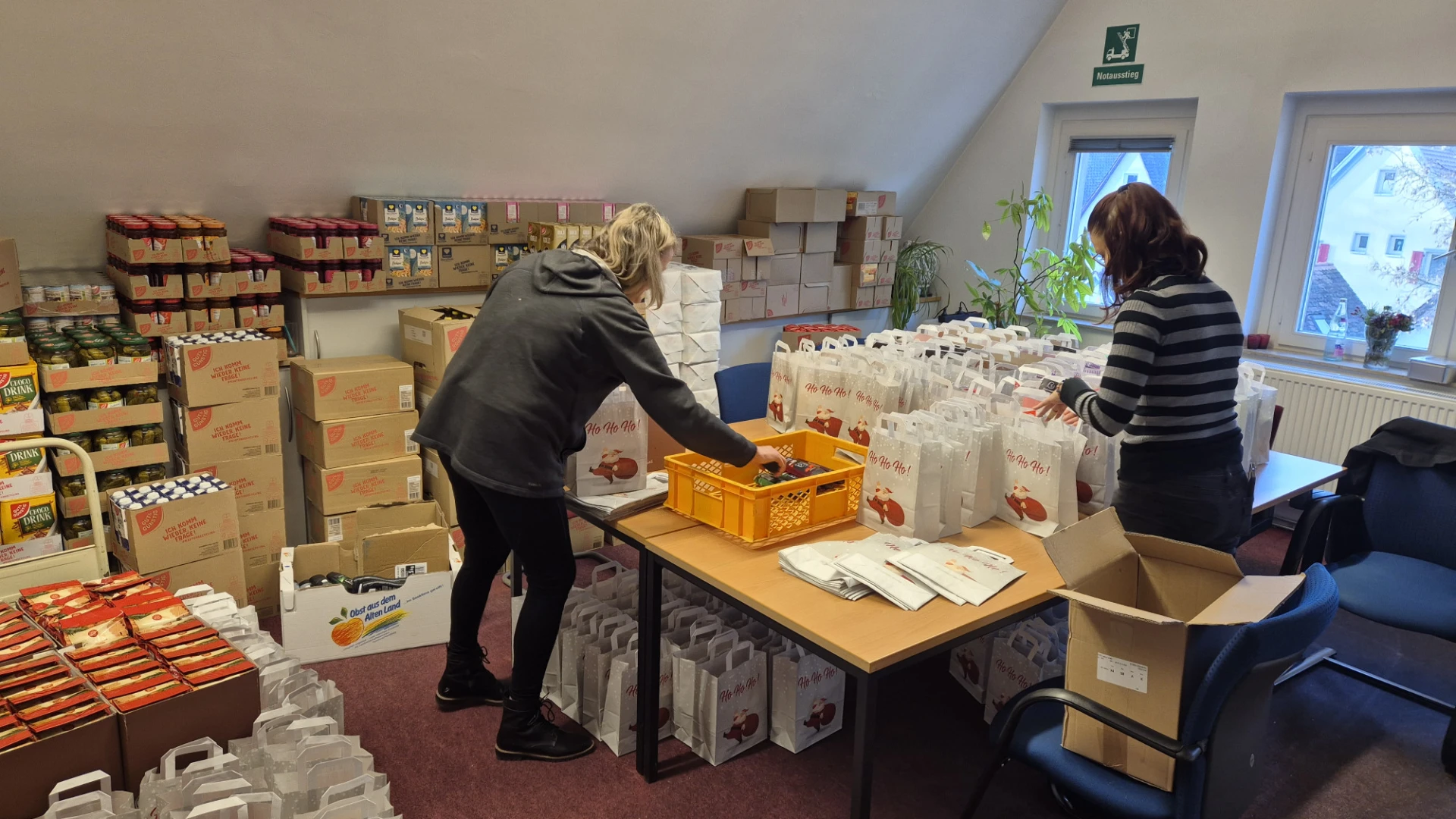 Zwei Personen packen Weihnachtsgeschenke in einem gro&szlig;en Lagerraum mit Regalen voller Kartons und P&auml;ckchen