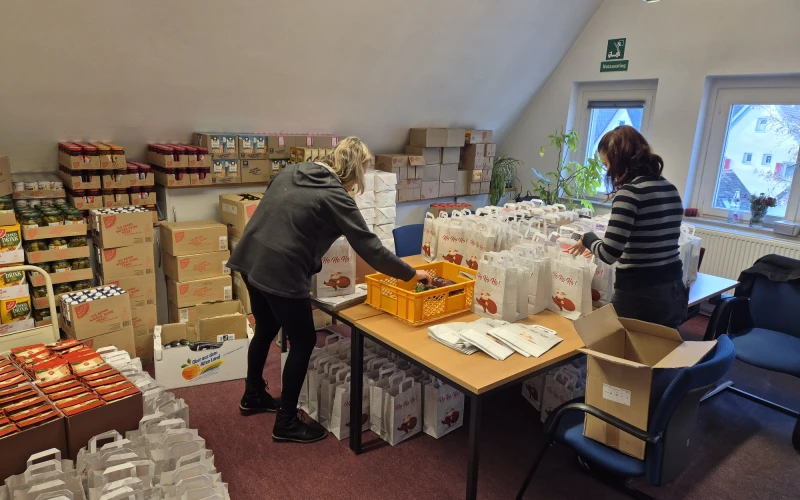Zwei Personen packen Weihnachtsgeschenke in einem gro&szlig;en Lagerraum mit Regalen voller Kartons und P&auml;ckchen