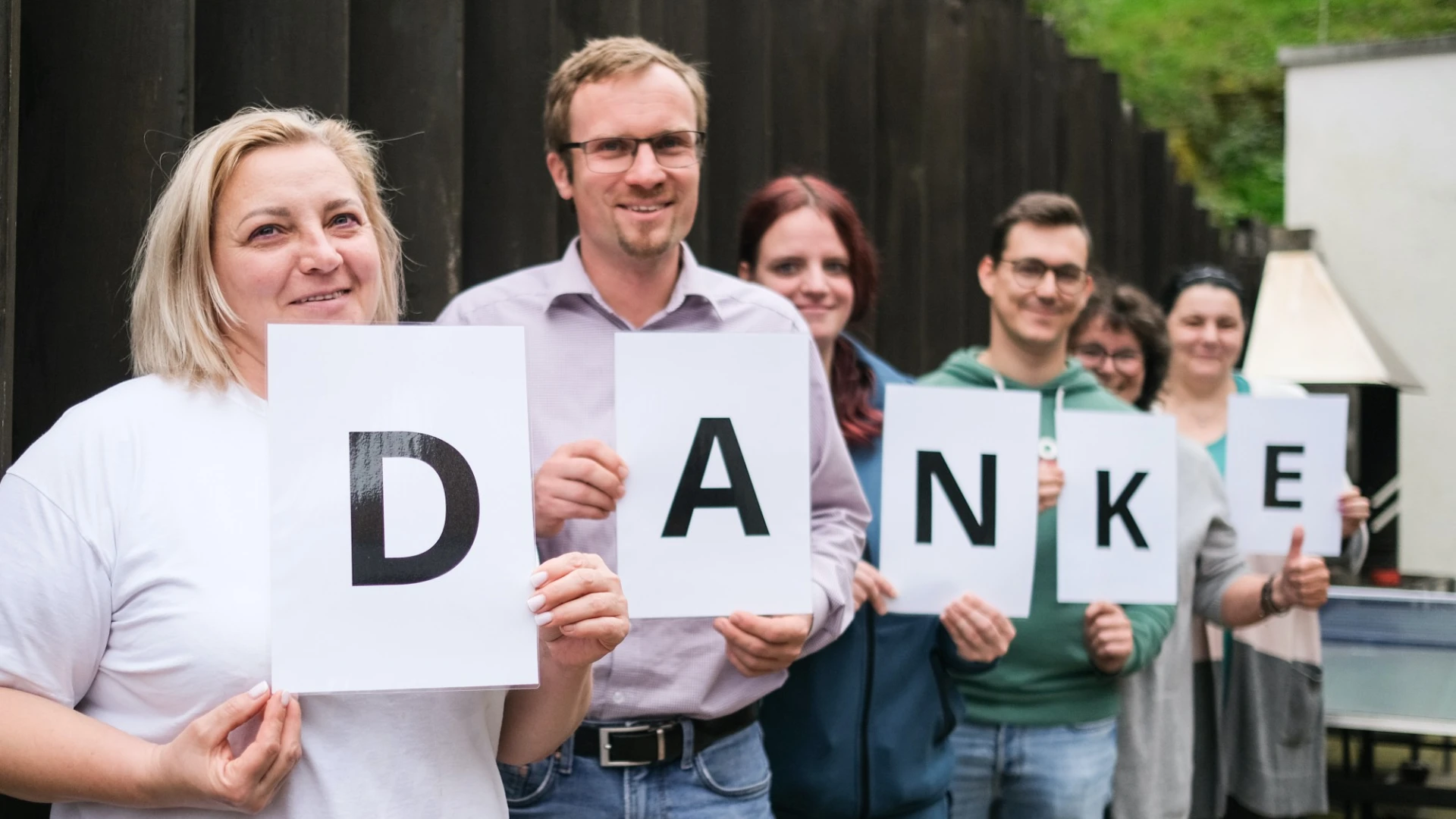 Sechs Personen stehen nebeneinander und halten wei&szlig;e Bl&auml;tter mit den Buchstaben von "DANKE" in den H&auml;nden, sie l&auml;cheln freundlich und tragen l&auml;ssige B&uuml;rokleidung vor einem dunklen Hintergrund.