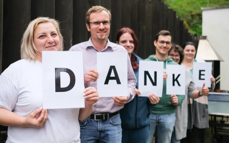 Sechs Personen stehen nebeneinander und halten wei&szlig;e Bl&auml;tter mit den Buchstaben von "DANKE" in den H&auml;nden, sie l&auml;cheln freundlich und tragen l&auml;ssige B&uuml;rokleidung vor einem dunklen Hintergrund.