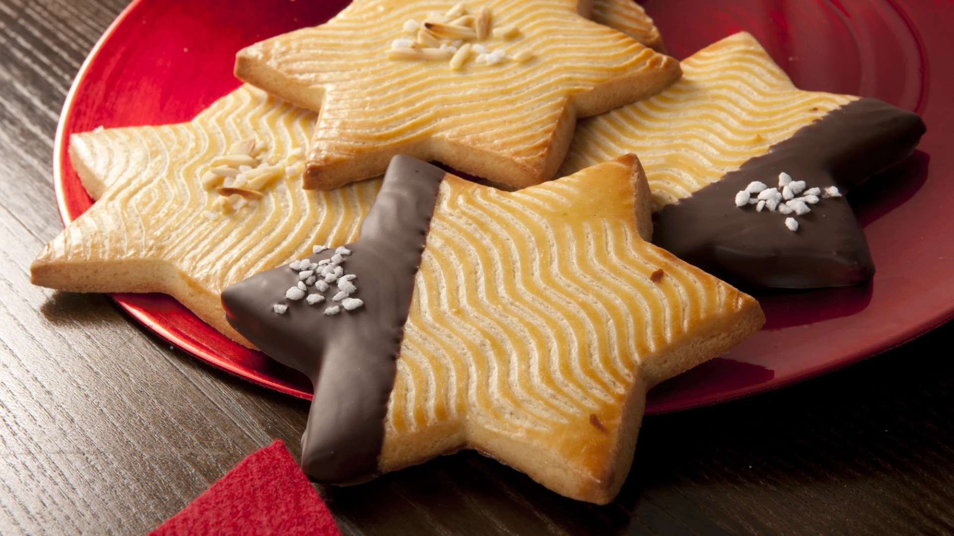 Sternf&ouml;rmige Weihnachtspl&auml;tzchen auf einem roten Teller, daneben eine rote Serviette auf einem dunklen Holzuntergrund. Die Pl&auml;tzchen haben eine goldbraune Farbe und eine wellenf&ouml;rmiges Muster auf der Oberseite. Teilweise sind sie mit Schokolade und Zuckerstreuseln verziert, teilweise mit Mandelsplitter und Zucker.