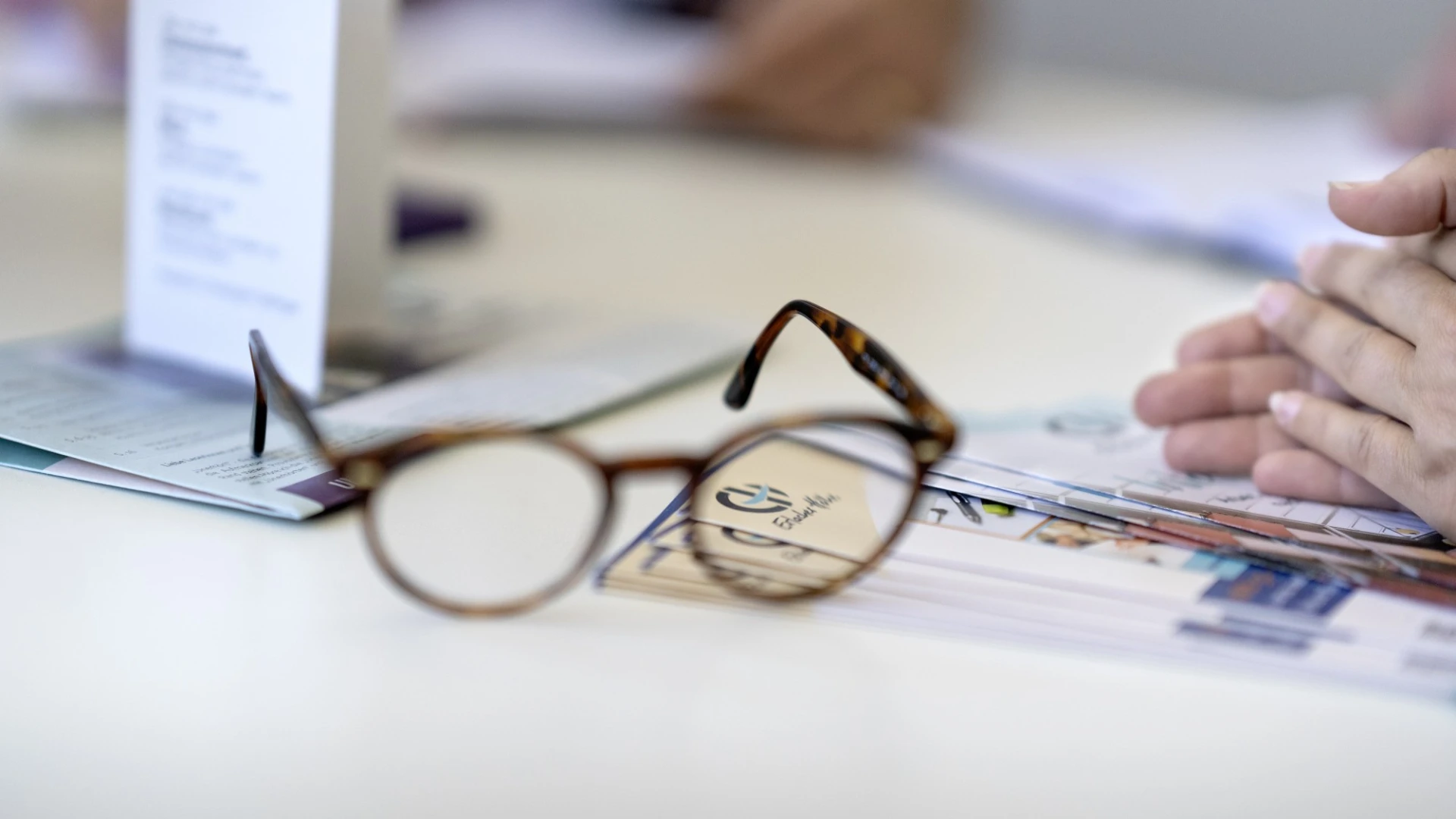 Auf einem Tisch liegt eine Brille auf Zeitung und Prospekten. Im Hintergrund sind unscharf H&auml;nde einer Person zu erkennen. Auf der rechten Seite sitzt ebenfalls eine Personen, deren H&auml;nde gefaltet auf dem Tisch liegen.