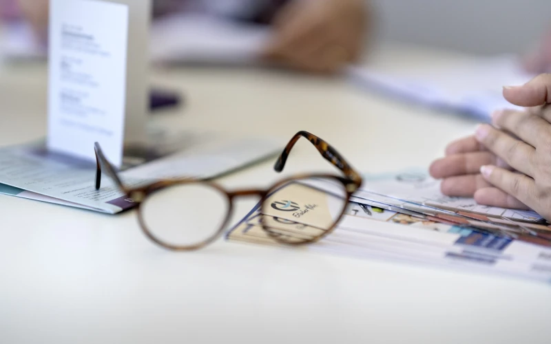 Auf einem Tisch liegt eine Brille auf Zeitung und Prospekten. Im Hintergrund sind unscharf H&auml;nde einer Person zu erkennen. Auf der rechten Seite sitzt ebenfalls eine Personen, deren H&auml;nde gefaltet auf dem Tisch liegen.