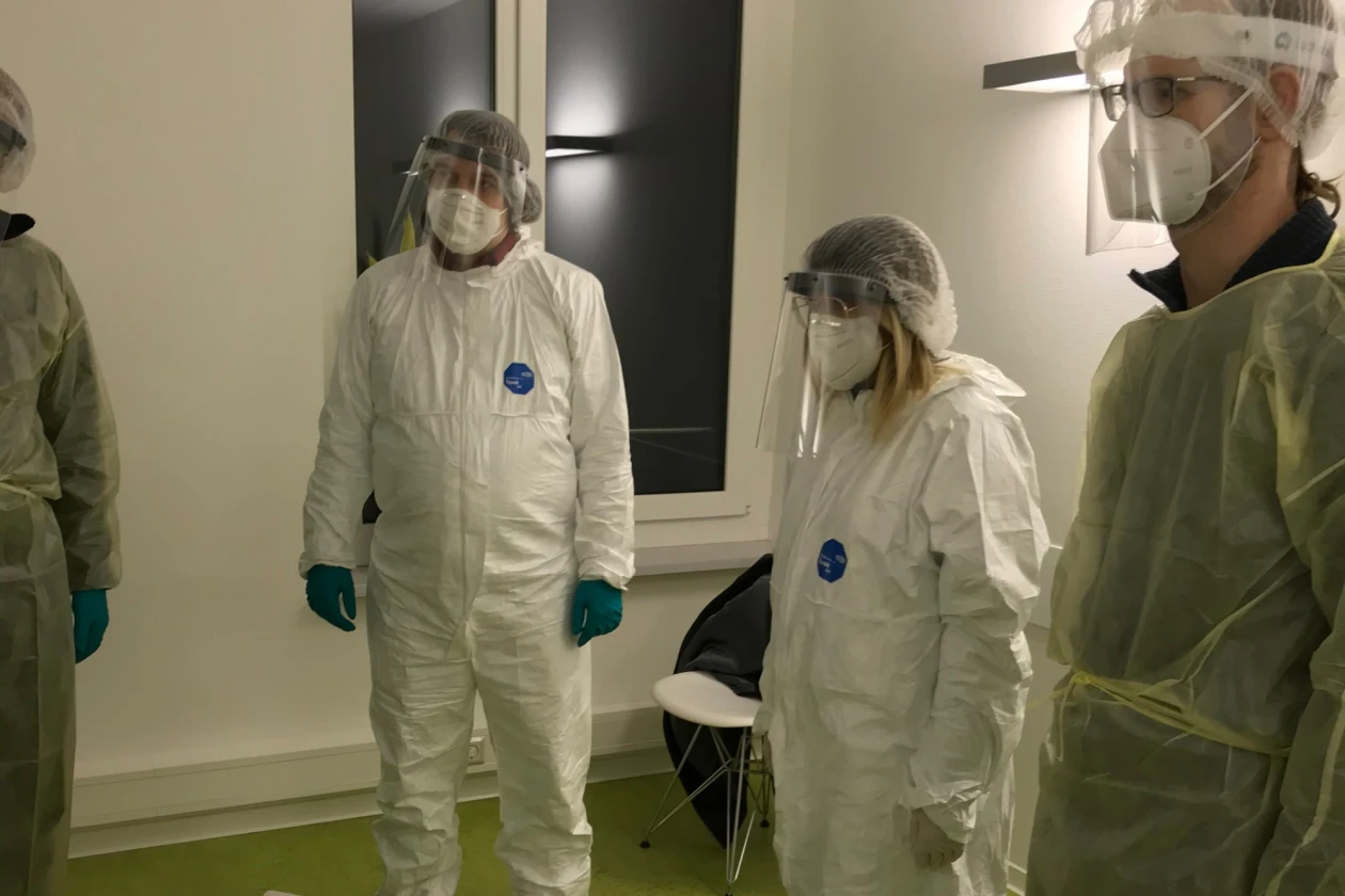 Vier Personen in vollst&auml;ndiger Schutzkleidung mit Gesichtsvisier, Mund-Nasenschutz und Handschuhen stehen in einem Raum mit Fenster und einem Stuhl.