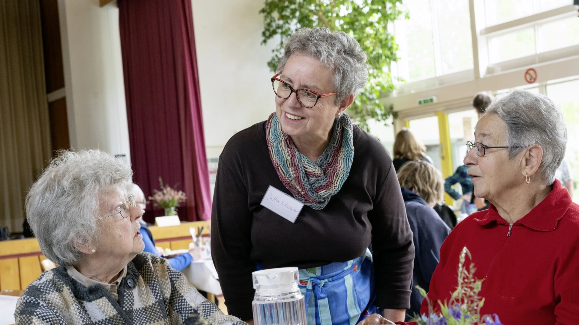Drei Frauen befinden sich an einem eingedeckten Tisch mit Getr&auml;nken und Blumendekoration. Eine Frau mit einem Namensschild beugt sich leicht zu den beiden sitzenden Frauen. Im Hintergrund ist eine B&uuml;hne, ein Fenster sowie weitere Personen zu sehen.