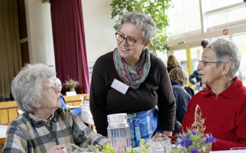 Drei Frauen befinden sich an einem eingedeckten Tisch mit Getr&auml;nken und Blumendekoration. Eine Frau mit einem Namensschild beugt sich leicht zu den beiden sitzenden Frauen. Im Hintergrund ist eine B&uuml;hne, ein Fenster sowie weitere Personen zu sehen.