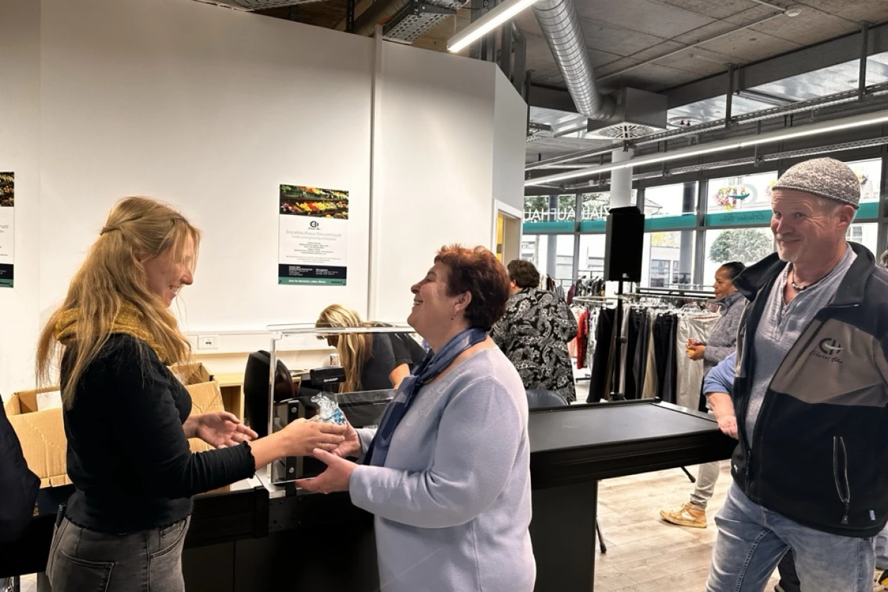 Zwei Personen unterhalten sich in einem hellen Ladenraum an der Verkaufstheke, eine dritte Person steht im Hintergrund. Die Umgebung sieht wie ein moderner Industrielook oder eine Begnungsst&auml;tte aus, mit Exponaten an der Wand.