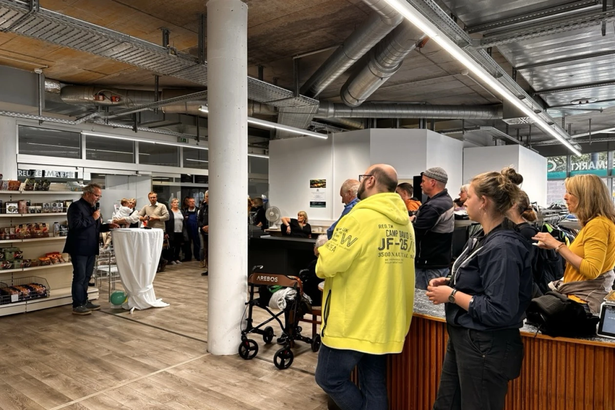 Mehrere Personen stehen in einem Raum eines Ladengesch&auml;fts mit wei&szlig;en S&auml;ulen, Holzboden und sichtbarer Deckeninstallation. Eine Gruppe steht an einer Theke, w&auml;hrend eine andere Menschengruppe im Hintergrund stehen.
