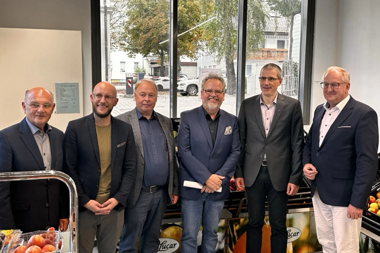 Sechs M&auml;nner in Business-Outfit stehen in einem Ladengesch&auml;ft, im Hintergrund gro&szlig;e Fenster und ein Blick nach drau&szlig;en. Sie posieren f&uuml;r ein Gruppenfoto in professioneller Atmosph&auml;re, mit einigen Gegenst&auml;nden im Vordergrund.