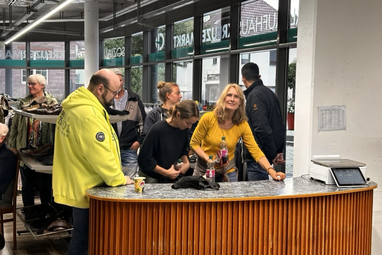 Mehrere Personen stehen an einem Holztresen in einem Ladengesch&auml;ft, gekleidet in verschiedene Farben wie Gelb, Schwarz und helle T&ouml;ne. Eine Person in einer gelben Jacke steht links am Tresen, w&auml;hrend Personen miteinander interagieren.