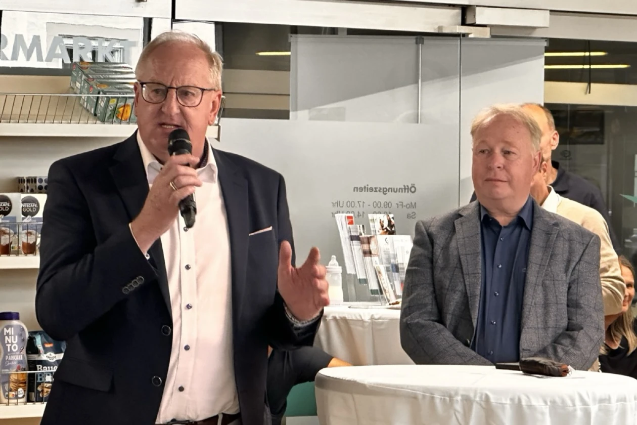 Zwei Herren in Anz&uuml;gen bei einer Veranstaltung. Der linke Mann spricht in ein Mikrofon und gestikuliert, w&auml;hrend der rechte Mann daneben steht. Sie befinden sich in einem Ladenraum mit Regalen und Mobiliar im Hintergrund.