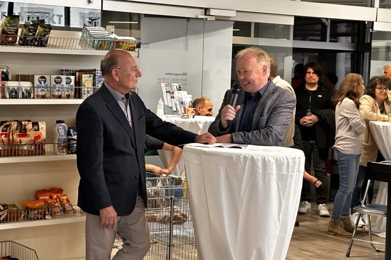 Zwei M&auml;nner in Businesskleidung unterhalten sich an einem wei&szlig;en Stehtisch, umgeben von Regalen mit Produkten und Personen im Hintergrund eines Ladengesch&auml;fts.