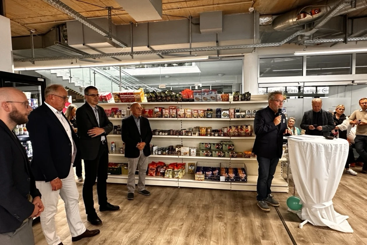 Mehrere M&auml;nner in Business-Outfit stehen vor einem Regal mit Lebensmittelprodukten und h&ouml;ren aufmerksam zu, w&auml;hrend ein Mann am Rednerpult mit einem Mikrofon spricht.