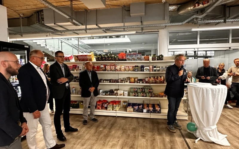 Mehrere M&auml;nner in Business-Outfit stehen vor einem Regal mit Lebensmittelprodukten und h&ouml;ren aufmerksam zu, w&auml;hrend ein Mann am Rednerpult mit einem Mikrofon spricht.