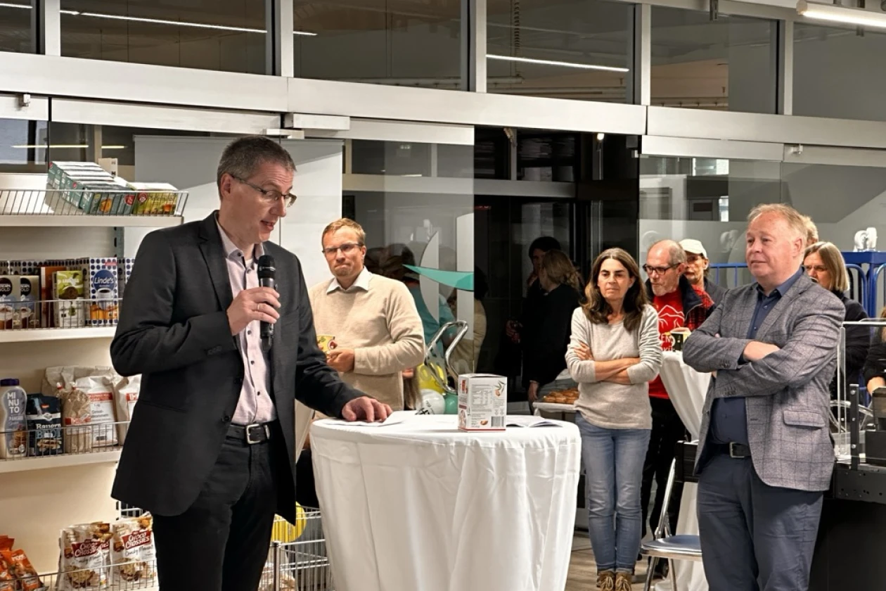 Ein Mann in schwarzem Anzug spricht mit einem Mikrofon bei einer Veranstaltung in einem Ladenraum, umgeben von mehreren Zuh&ouml;rern unterschiedlichen Alters, die aufmerksam zuh&ouml;ren. Im Hintergrund sind Regale mit Produkten und eine Theke mit wei&szlig;er Tischdecke zu sehen.