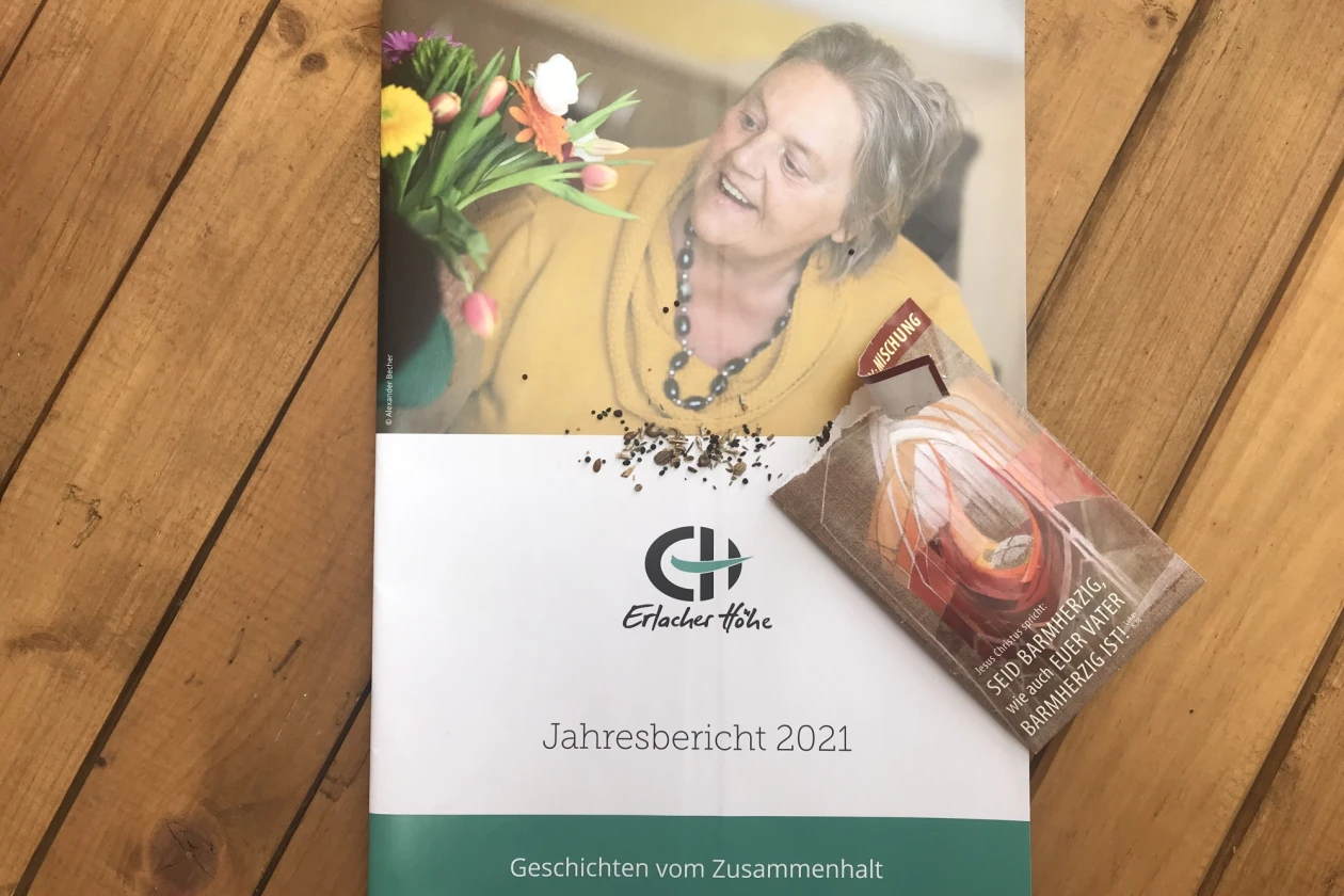 Zu sehen ist die Brosch&uuml;re "Jahresbericht 2021" der Erlacher H&ouml;he mit dem Titel "Geschichten vom Zusammenhalt liegt auf einem Holztisch. Daneben liegt eine kleine Samenpackung, aus der einige Samen auf das Heft gefallen sind.