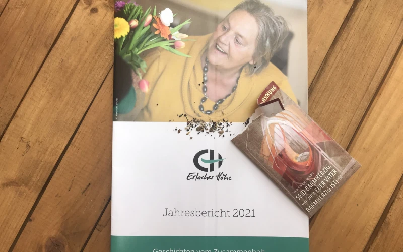 Zu sehen ist die Brosch&uuml;re "Jahresbericht 2021" der Erlacher H&ouml;he mit dem Titel "Geschichten vom Zusammenhalt liegt auf einem Holztisch. Daneben liegt eine kleine Samenpackung, aus der einige Samen auf das Heft gefallen sind.