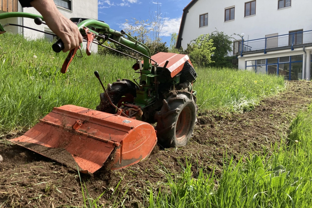 In einem Wiesenst&uuml;ck wird ein St&uuml;ck Erde mit einer Agria-Motofr&auml;se bearbeitet. Im Hintergrund sind zwei Geb&auml;ude zu sehen.