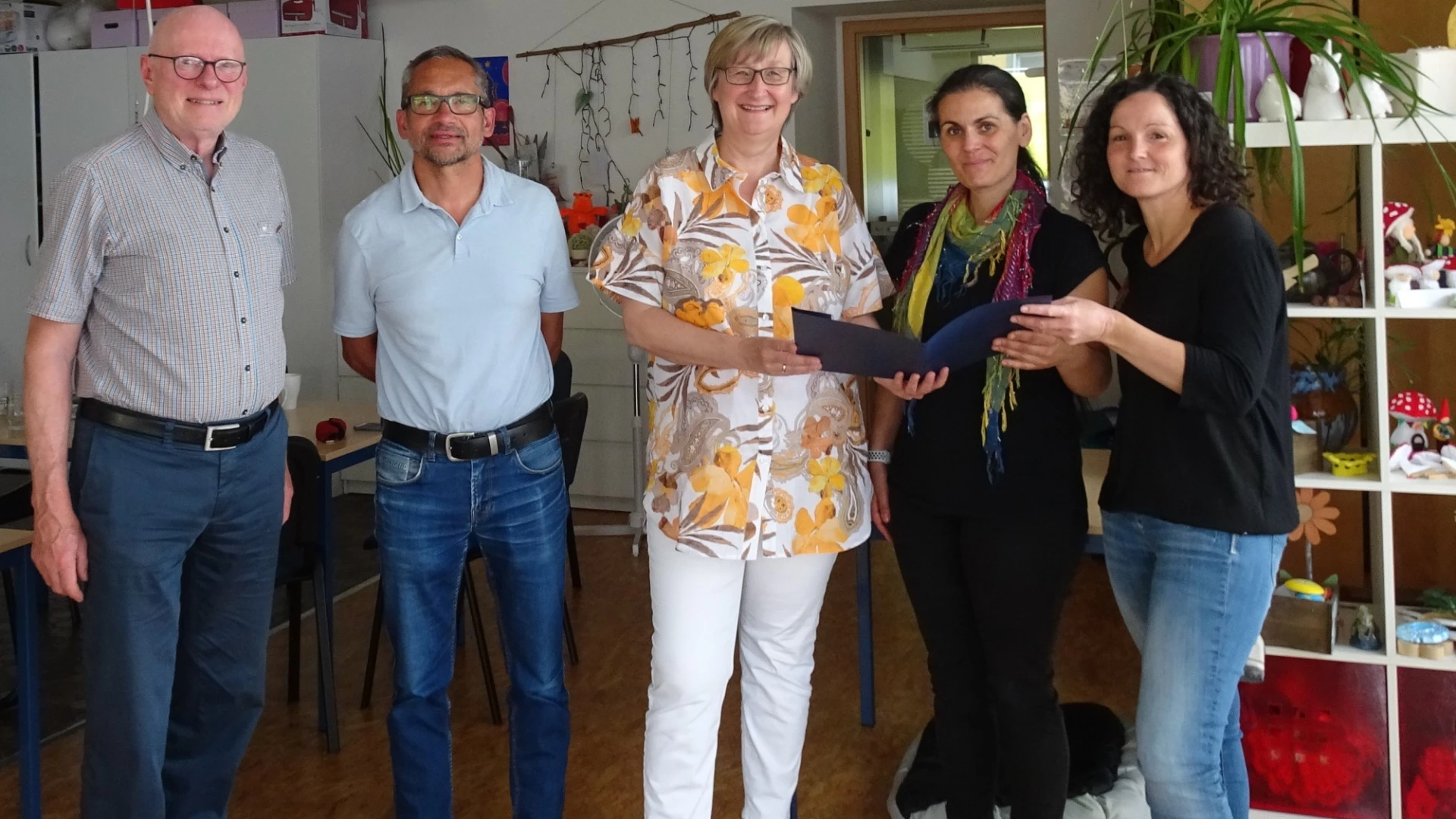 Zwei M&auml;nner und drei Frauen blicken freudig in die Kamera. Sie stehen in einem Raum mit Regal und Fenster im Hintergrund. Die Frauen halten ein Dokument in der Hand.