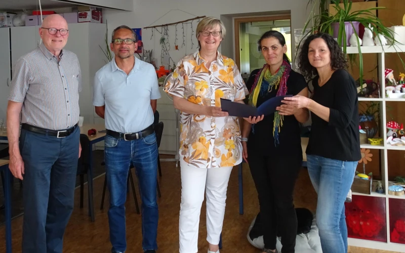 Zwei M&auml;nner und drei Frauen blicken freudig in die Kamera. Sie stehen in einem Raum mit Regal und Fenster im Hintergrund. Die Frauen halten ein Dokument in der Hand.