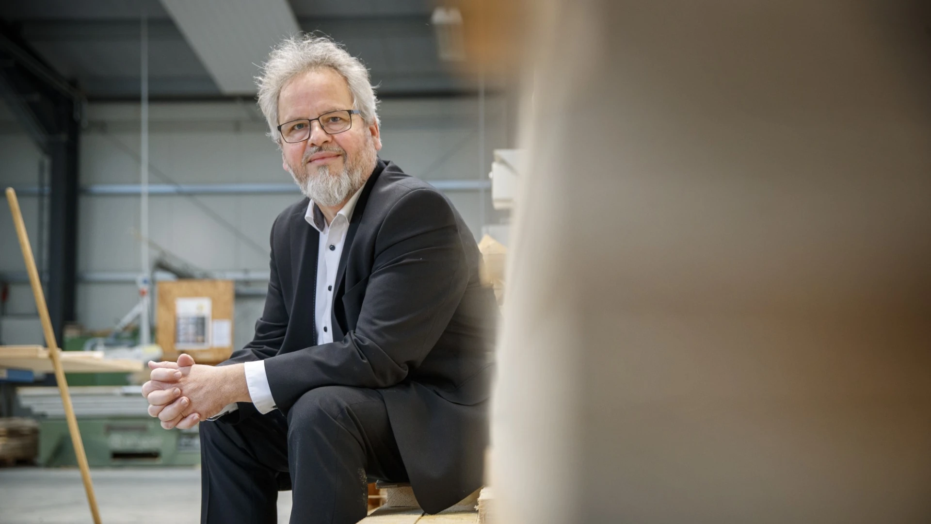Ein &auml;lterer Mann mit grauem Haar, Bart und Brille sitzt in einem schwarzen Anzug und wei&szlig;em Hemd auf einem Holzstapel in einer Lagerhalle.