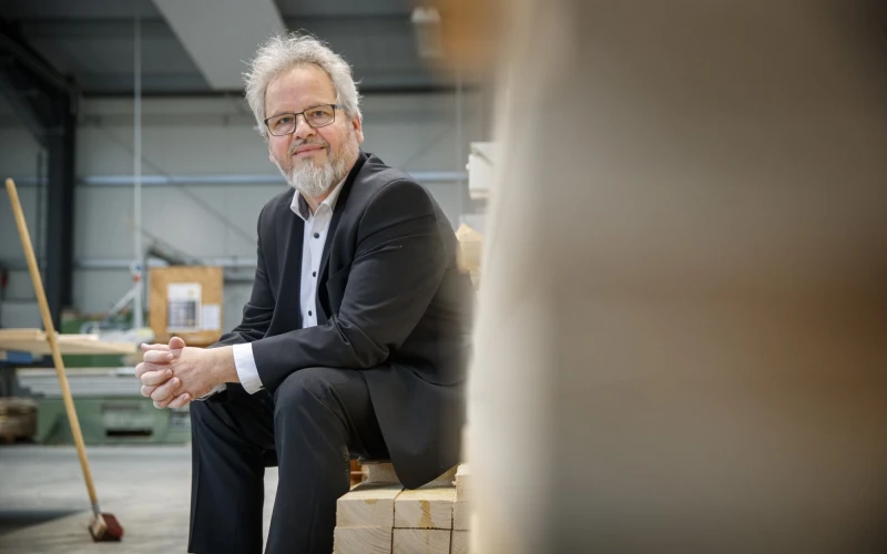 Ein &auml;lterer Mann mit grauem Haar, Bart und Brille sitzt in einem schwarzen Anzug und wei&szlig;em Hemd auf einem Holzstapel in einer Lagerhalle.
