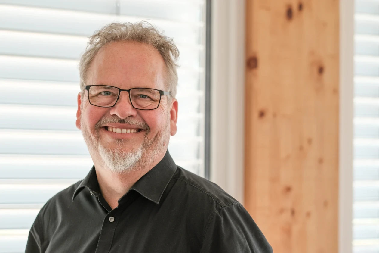Ein Mann mit grauen Haaren und Bart l&auml;chelt freundlich. Er hat eine Brille und tr&auml;gt ein schwarzes Hemd.