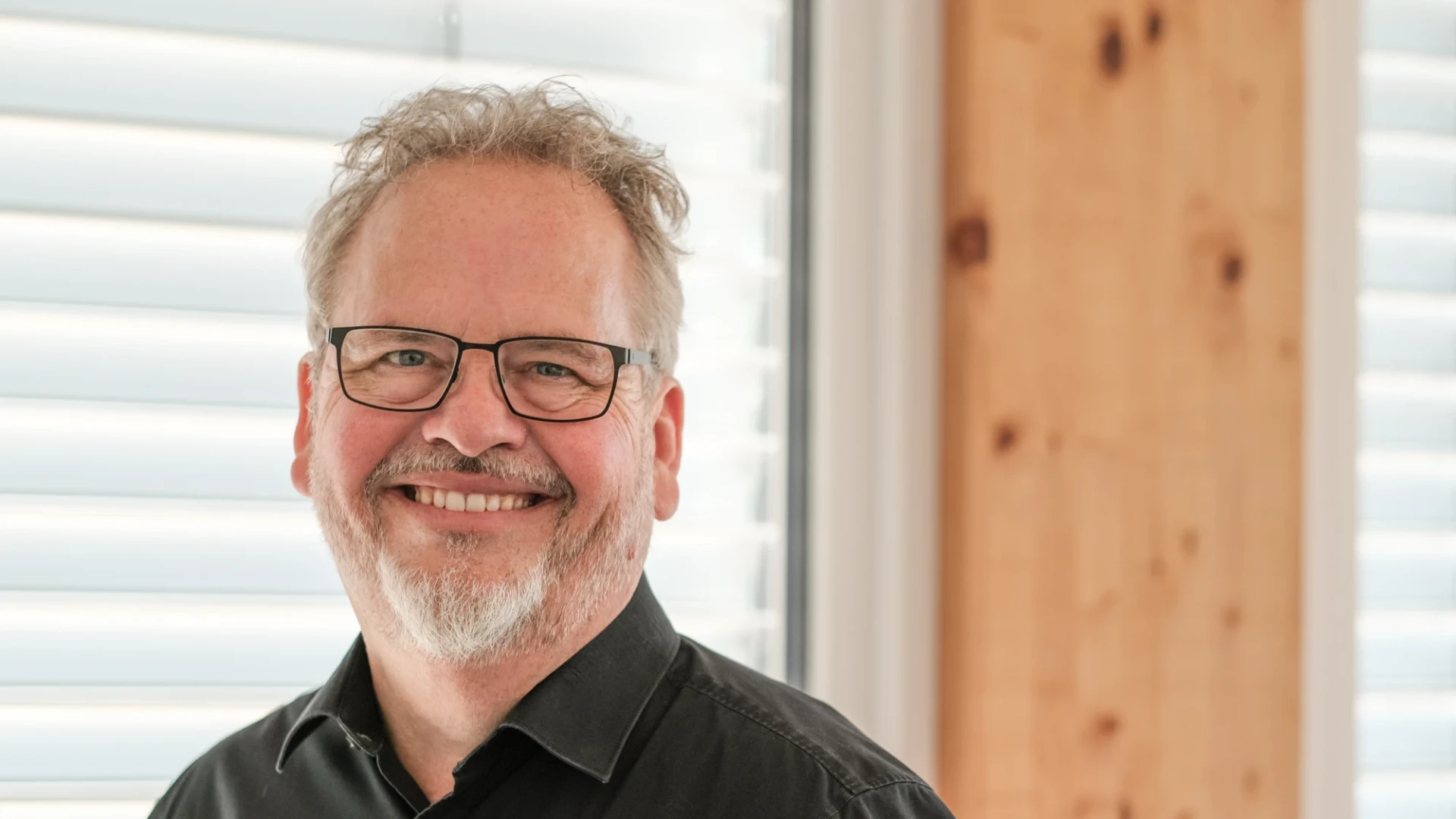 Ein Mann mit grauen Haaren und Bart l&auml;chelt freundlich. Er hat eine Brille und tr&auml;gt ein schwarzes Hemd.