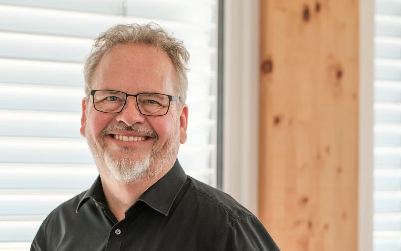 Ein Mann mit grauen Haaren und Bart l&auml;chelt freundlich. Er hat eine Brille und tr&auml;gt ein schwarzes Hemd.