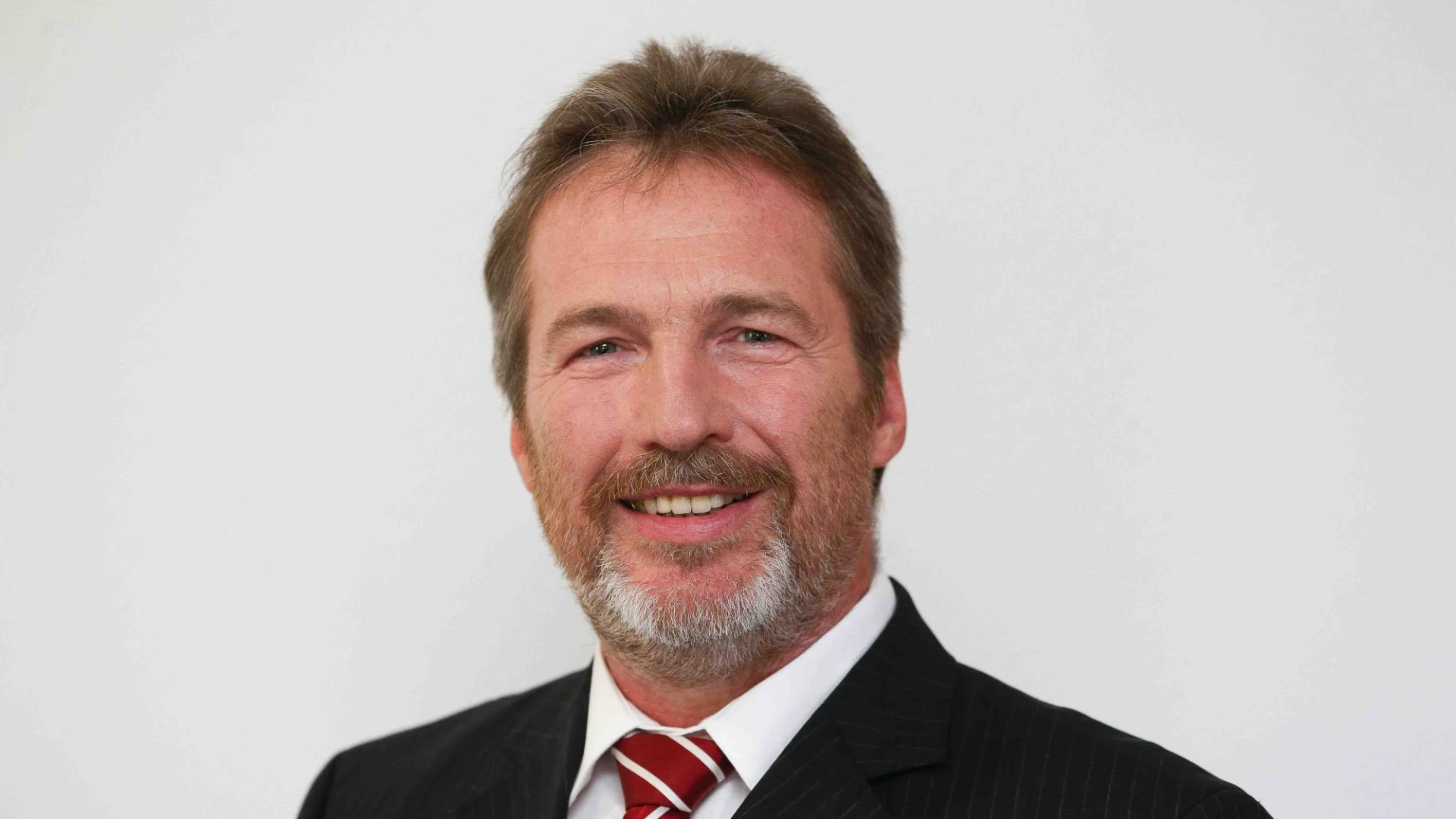 Ein Mann mit schwarzem Anzug, wei&szlig;em Hemd und rot-wei&szlig;er Krawatte ist zu sehen. Er l&auml;chelt direkt in die Kamera.