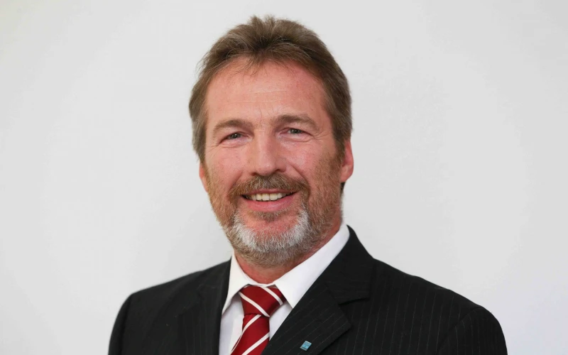 Ein Mann mit schwarzem Anzug, wei&szlig;em Hemd und rot-wei&szlig;er Krawatte ist zu sehen. Er l&auml;chelt direkt in die Kamera.