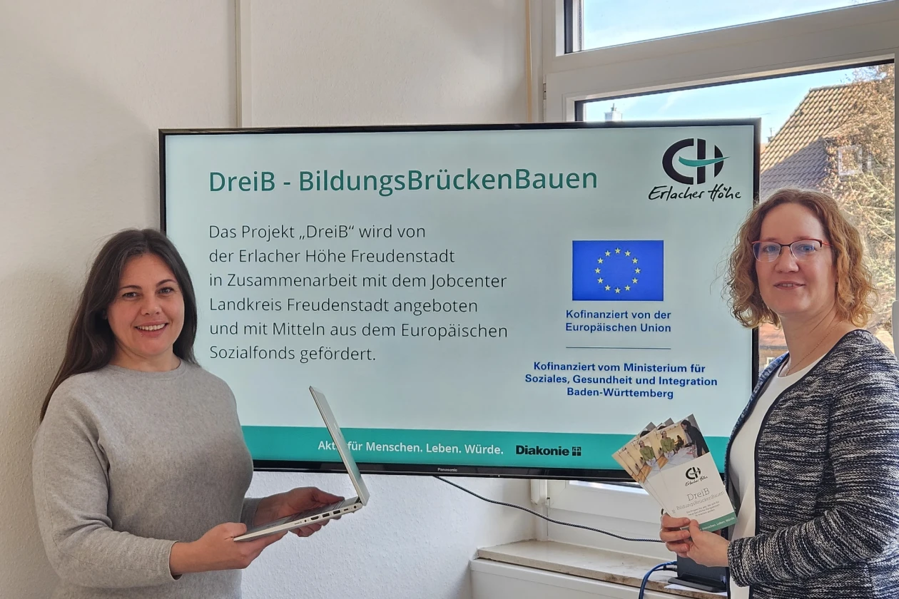 Vor einem gro&szlig;en Monitor stehen zwei junge Frauen. Die Frau links hat lange dunkle Haare und einen Laptop in der Hand. Die Frau rechts hat eine Brille und h&auml;lt Flyer in der Hand. Auf dem Monitor ist zu lesen "DreiB - BildungsBr&uuml;ckenBauen".