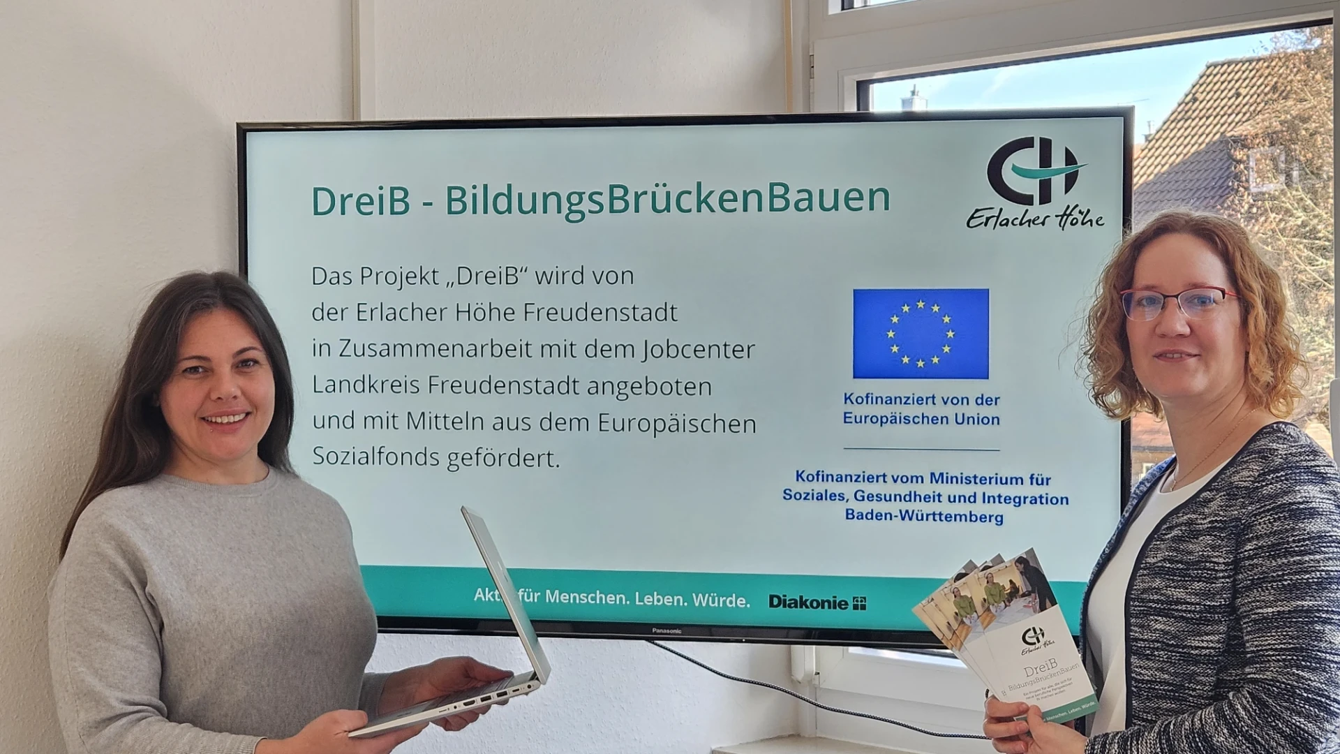 Vor einem gro&szlig;en Monitor stehen zwei junge Frauen. Die Frau links hat lange dunkle Haare und einen Laptop in der Hand. Die Frau rechts hat eine Brille und h&auml;lt Flyer in der Hand. Auf dem Monitor ist zu lesen "DreiB - BildungsBr&uuml;ckenBauen".