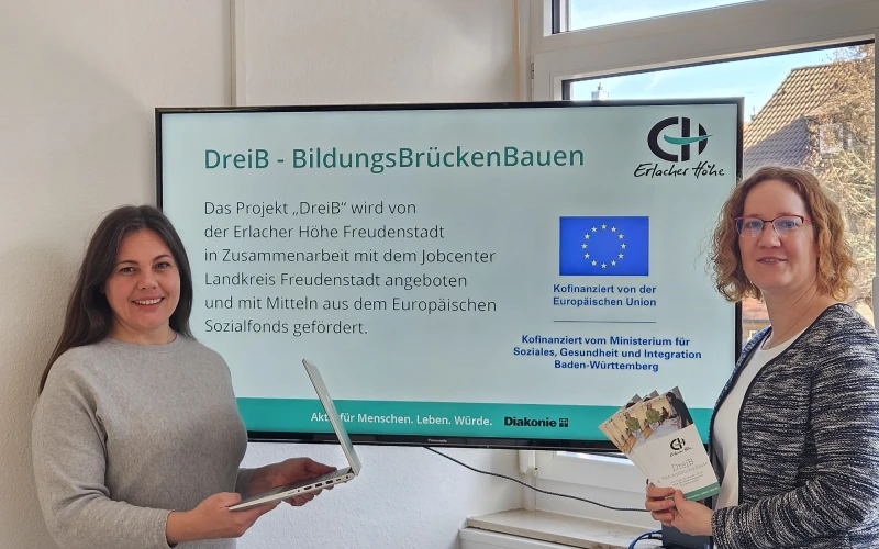 Vor einem gro&szlig;en Monitor stehen zwei junge Frauen. Die Frau links hat lange dunkle Haare und einen Laptop in der Hand. Die Frau rechts hat eine Brille und h&auml;lt Flyer in der Hand. Auf dem Monitor ist zu lesen "DreiB - BildungsBr&uuml;ckenBauen".