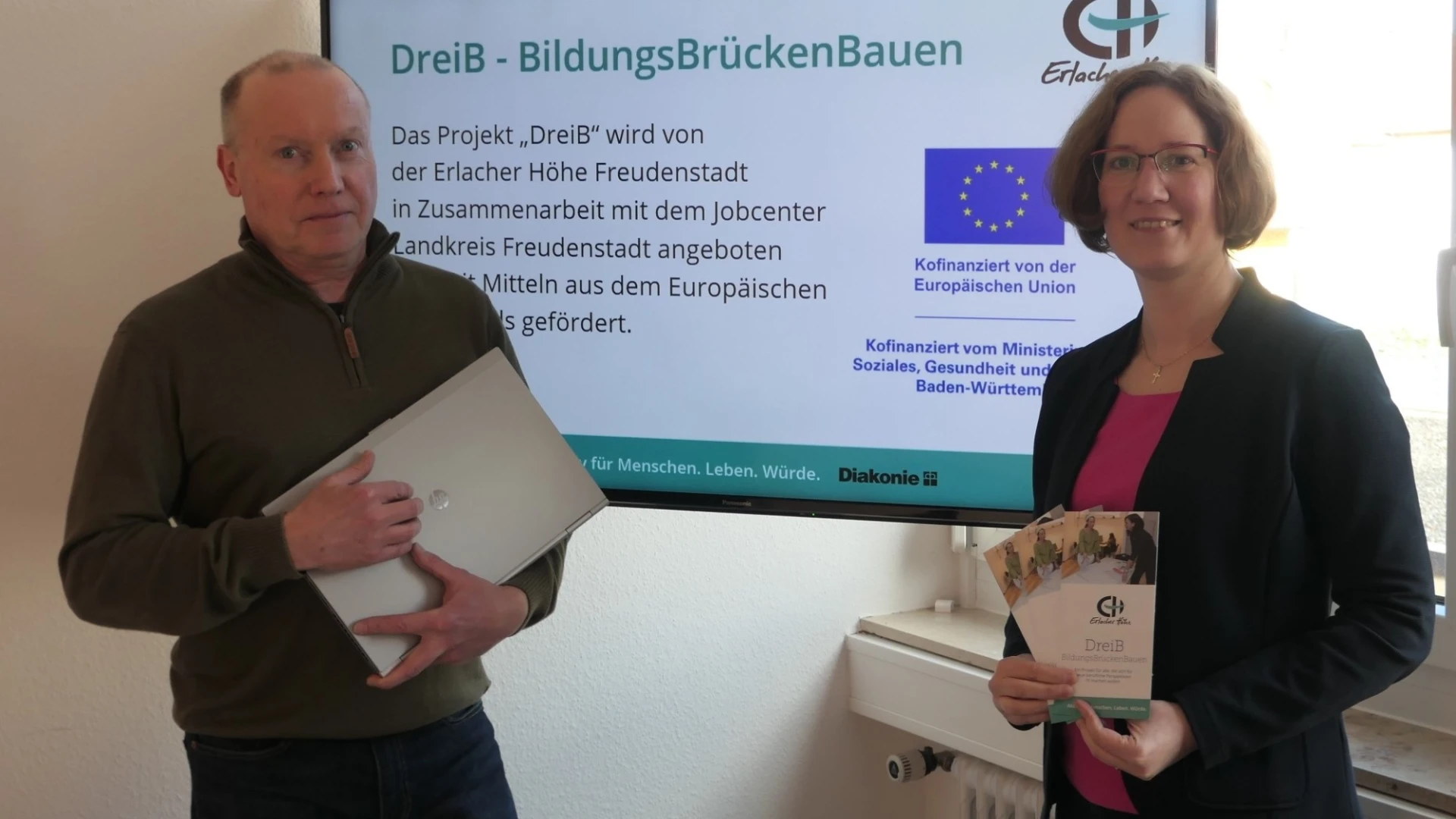 Eine Frau und ein Mann blicken in die Kamera und stehen vor einem Bildschirm mit einer Pr&auml;sentation zum BildungsBr&uuml;ckenBauen. Es scheint sich um eine Informationsveranstaltung zu handeln.