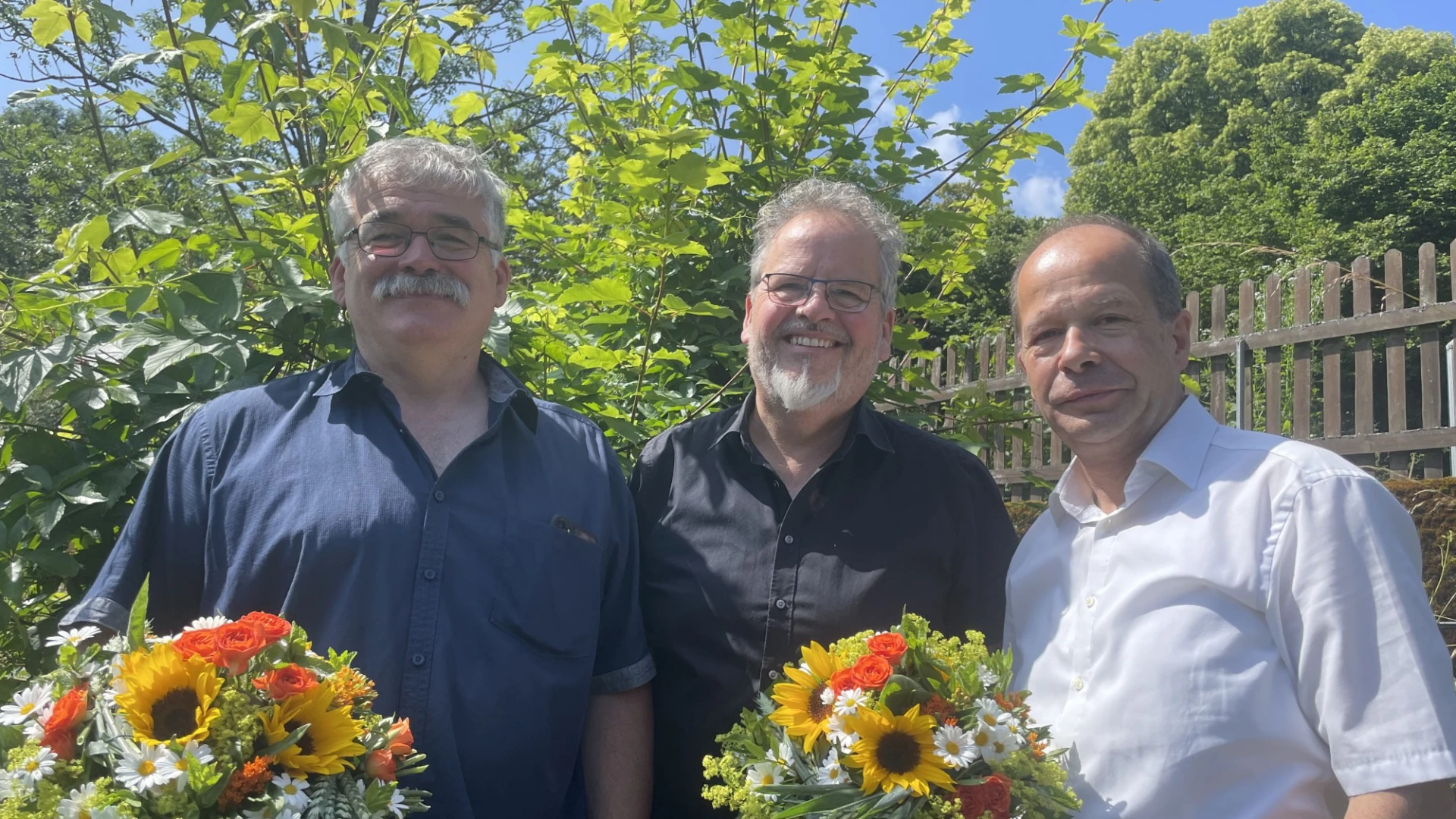 Drei M&auml;nner blicken freundlich in die Kamera. Zwei halten einen Blumenstrau&szlig;. Im Hintergrund sind gr&uuml;ne Hecken, B&auml;ume und ein Gartenzaun zu sehen.